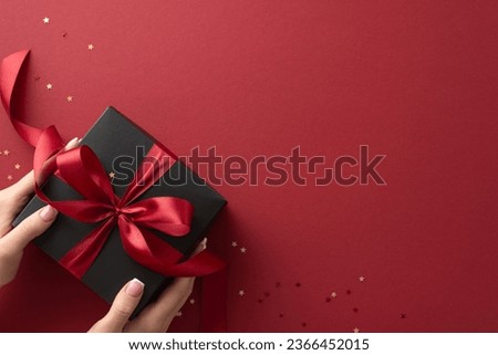 Festive vibe captured! First person top view of my hands caressing exquisite black gift box tied with a red ribbon amid golden star confetti on vibrant marsala backdrop. Perfect for holiday promotions
