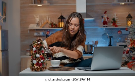 Festive Woman Talking On Video Call Using Laptop In Christmas Decorated Room. Young Adult Celebrating Traditional Holiday On Internet Comunication At Home Pouring Cup Of Tea From Kettle