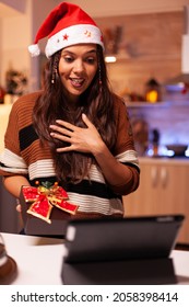 Festive Young Adult Holding Christmas Gift On Internet Conference Video Call With Friends And Family Using Tablet. Caucasian Cheerful Woman Showing Present Box With Bow And Ribbon