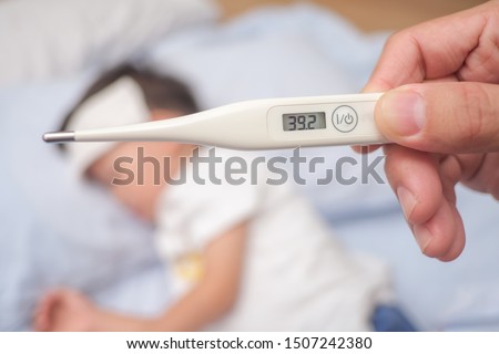 Fever, Close-up medical thermometer, Parent / Father measuring temperature of his ill kid, Asian 3 - 4 years old toddler boy gets high fever lying on bed with cold compress on forehead to cool a fever