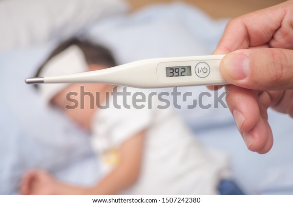 Fever, Close-up medical thermometer, Parent /
Father measuring temperature of his ill kid, Asian 3 - 4 years old
toddler boy gets high fever lying on bed with cold compress on
forehead to cool a fever