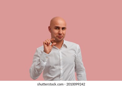 Few inches, centimeter. Funny bald man with bristle in white shirt, showing a little bit gesture, asks for something, assuring that it is just a little bit. Isolated over pink background