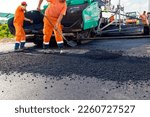 Few workers are using shovels to level hot tarmac on prepared ground, in background is machine for laying asphalt, spreading layer.