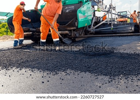 Few workers are using shovels to level hot tarmac on prepared ground, in background is machine for laying asphalt, spreading layer.