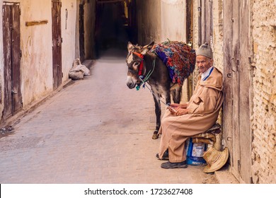 Fez, Morroco - October 31, 2015. An Elderly Man, Wearing Traditional Moroccan Clothing, Sits With His Pack Donkey Outside The Local Market.