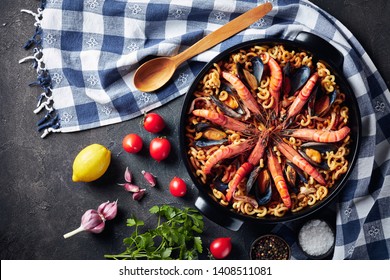 Fideua, A Paella Like Spanish Dish With Special Pasta, King Prawns, White Fish Meat, Calamari, Mussels In A Black Pan On A Concrete Table With Ingredients, View From Above, Flatlay, Empty Space