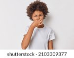 Fie something stinks. Displeased curly haired woman covers nose smells something unpleasant has disgusting expression wears casual t shirt isolated over white background. Bad smell coming from kitchen