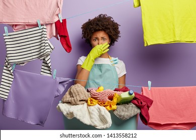 Fie, What Stench! Shocked Dissatisfied Ethnic Female Housekeeper Closes Nose, Smells Dirty Laundry From Basket, Busy With Washing Clothes During Weekend, Wears Green Rubber Gloves And Apron.