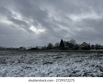 field covered in a light dusting of snow under a heavily overcast sky.
Several houses and buildings are visible in the distance, nestled among bare trees and evergreens.