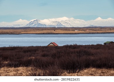 A field of dark bushes frames a small cabin with a red roof overlooking a wide body of water in Iceland. In the background stretches a rural plain dominated by imposing, completely snow-covered mountain peaks under a clear sky.