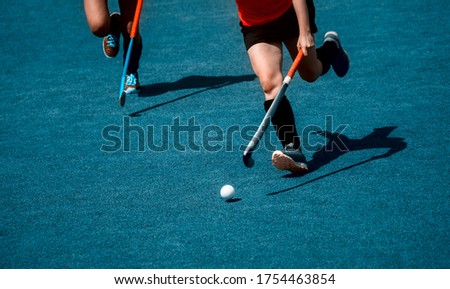 Field hockey player, in possesion of the ball, running over an astroturf pitch, looking for a team mate to pass to. Blue color filter