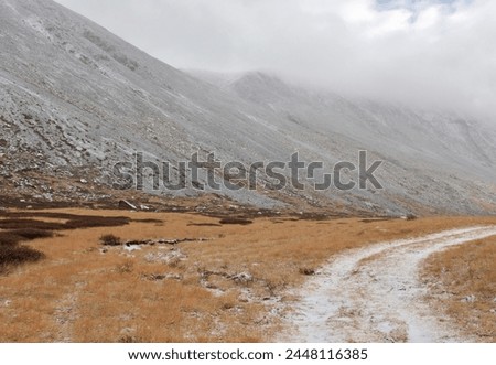 A field road covered with snow turns right along a high mountain range on a cloudy autumn morning.