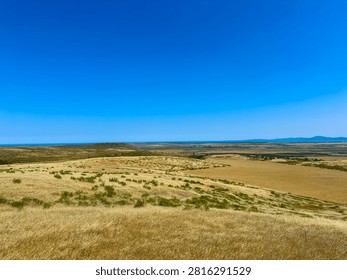 Fields stretch across the view with dry grass and sparse plants. In the distance, a body of water reflects the blue sky. The scene captures open space and bright sunlight.