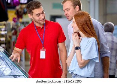 Fiendly Consultant Help Customers Couple In Store, Make Choice, Buy Smartphone, Talking, Having Discussion. Male Consultant In Red Uniform Working With Clients Talk About Models Options - Shutterstock ID 2116195205