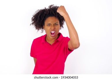 Fierce young beautiful African American woman wearing pink t-shirt against white wall holding fist in front as if is ready for fight or challenge, screaming and having aggressive expression on face.