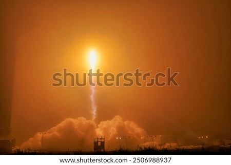 A fiery explosion lights up the night sky over a city, creating a dramatic scene. Smoke and flames rise high, illuminating the buildings and vehicles belo. Elements of this image furnished by NASA