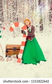 fiery red-haired woman in a ball green dress with a red leather belt in the costume of dwarf assistant Santa Claus in the winter forest with huge candy, a chest of gifts, the concept of the new year