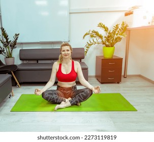 A fifty-year-old athletic woman in a red tank top sits in a lotus position on a green sports mat in her office. Workout in the office. Adult athletic woman - Powered by Shutterstock