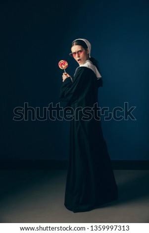 Fight for your rights. Medieval young woman as a nun in vintage clothing, white mutch and red sunglasses eating sweets and standing on dark blue background. Concept of comparison of eras.