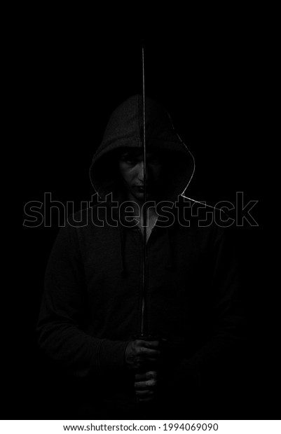 A fighter in black clothes with a hood on his\
head holds the Japanese sword katana in the face dividing it into\
two halves on a black\
background