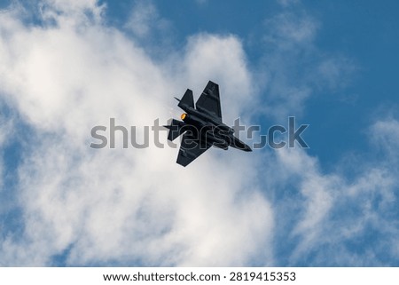 A fighter jet flies through clouds with its afterburner visible against a blue sky.