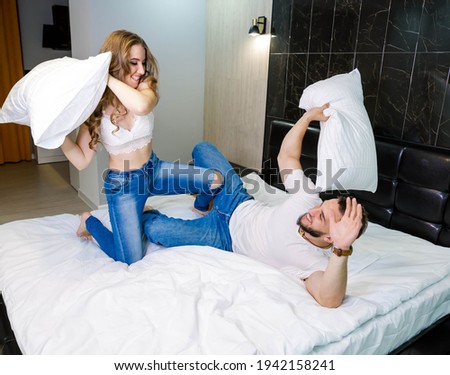 Fighting with pillows. Happy young european couple having fun. Playful woman beating scared boyfriend with pillow on bed. Smiling man and woman fooling around together at home, good morning.