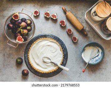 Figs tart preparation with fruits, bowls, cheesecake cream, wooden rolling pin and kitchen utensils at grey concrete table. Baking at home. Top view.