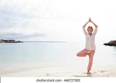 Figure Of Mature Beautiful Woman In Tree Yoga Position, On Shore Of Inviting, Calm Blue Sea Destination White Sand Beach, Sunny Holiday, Outdoors. Healthy Travel Fitness Lifestyle.