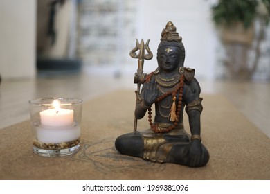 
A Figurine Of God Shiva And A Burning Candle In A Glass On A Yoga Mat With A Blurred Background