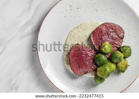 The filet mignon steak is cut in half and lies on a mustard sauce with mustard seeds. Nearby lies the Brussels capstsa boiled. Food lies on a light ceramic plate on a marble background.