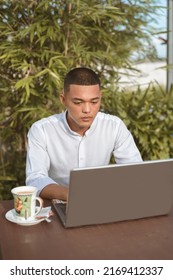 A Filipino Male Analyst Busy Reviewing The Data On His Laptop While Working Remotely At An Outdoor Cafe, Coffee Shop, Or Restaurant. Modern Freelance Professional Lifestyle.