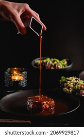 Fillet Steak On The Dark Background On The Table In Restaurant Fine Dining Dinner, Man Hand Sauce Pouring Over The Meat, Tasty Food Around , Romantic Atmosphere With Candle