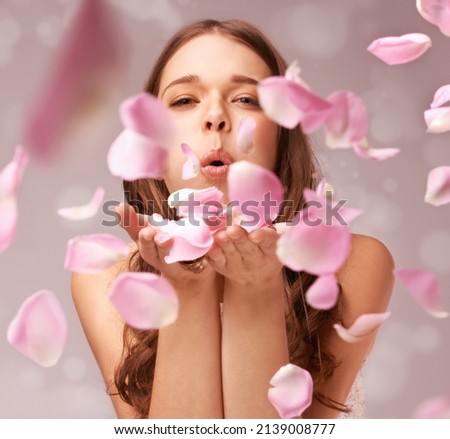 Filling the air with a whimsical scent. Gorgeous young woman blowing away a handful of rose petals on a pink background.