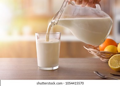 Filling glass of milk with jug for breakfast on brown wooden table in rustic kitchen. Horizontal composition. Front view - Shutterstock ID 1054415570