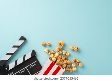 Film debut concept. Top view of moviemaker's clapperboard, and tasty striped popcorn container on soft blue backdrop with room for your message or advertisement