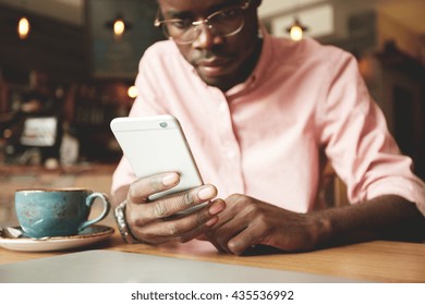 Film Effect. Handsome African Student In Shirt And Glasses Using Mobile Phone, Looking At The Screen With Serious And Concentrated Expression While Having Coffee At A Cafe After Classes At University