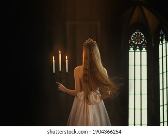 Film grain added. medieval girl princess walks in dark gothic room. Woman queen holding candlestick with burning candles in hand. Dress with open back, long loose blonde hair flying in motion. Go away