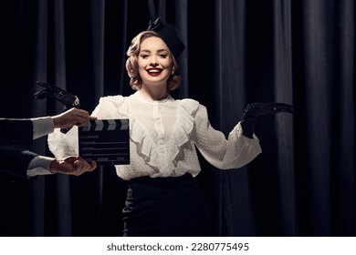Film shooting. Charming actress wearing vintage clothes and posing at camera and smiling over dark background. Concept of old films, actress, beauty, fashion, old films, actress, retro, vintage