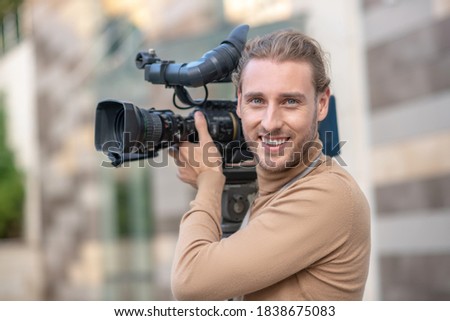 Filming process. Smiling fair-haired operator standing with camera on his shoulder outside