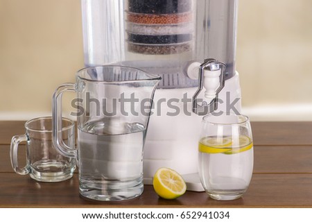Filter system of water purifier with two glasses of water one filled until middle with a lemon inside and an empty pitcher on wooden table