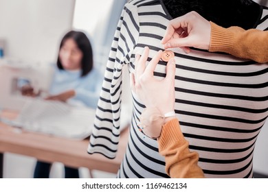 Final Steps. Close Up Of Skilled Young Seamstress Using A Needle And Sewing A Button On Dress While Her Coworker Using Sewing Machine