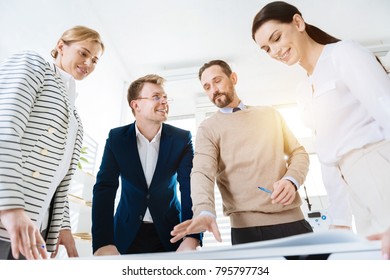 Final variant. Low angle of attractive jolly four  colleagues standing around table  while discussing important topic and grinning 