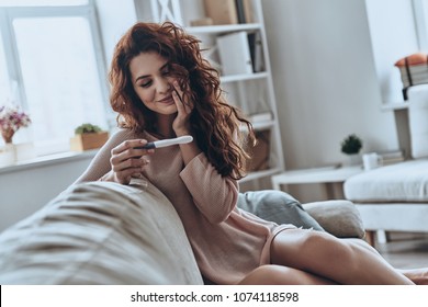 Finally pregnant. Attractive young women looking at pregnancy test and smiling while sitting on the sofa at home