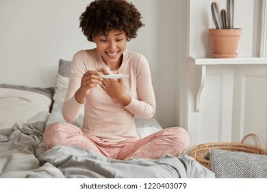 Finally Pregnant. Happy African American Woman Cant Believe In Positive Pregnancy Test Result, Looks Positively, Sits Crossed Legs, Wears Pyjamas, Enjoys Domestic Atmosphere. Women, Fertility Concept