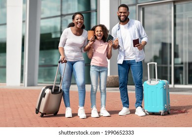 Finally Vacation. Cheerful positive black family of three people holding passports with tickets and shaking clenched fists, making winner gesture, travelling together standing near airport terminal - Shutterstock ID 1990652333