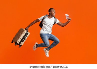 Finally Vacation. Young Happy Black Man With Suitcase, Backpack, Passport And Flight Tickets Jumping Up Orange Studio Background. Excited African American Guy Running Towards Copy Space