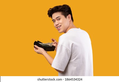 Finance, Investment And Money Saving. Portrait of excited asian young man holding and showing wallet full of dollars, free space, isolated over orange studio background. Greedy guy got salary