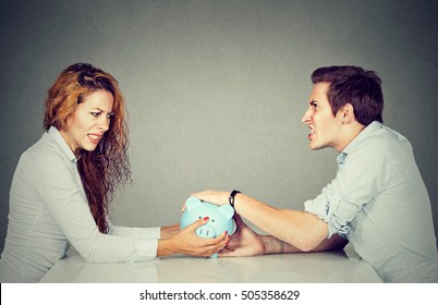 Finances In Divorce Concept. Wife And Husband Can Not Make Settlement Holding Piggy Bank Sitting At Table Looking At Each Other With Hatred