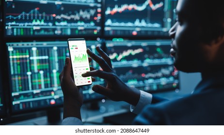 Financial Analyst Using Smartphone with Real-Time Stocks, Commodities and Market Charts. Working on Multi-Monitor Workstation with Stock Charts. Businessman Works in Investment Bank in the Evening.