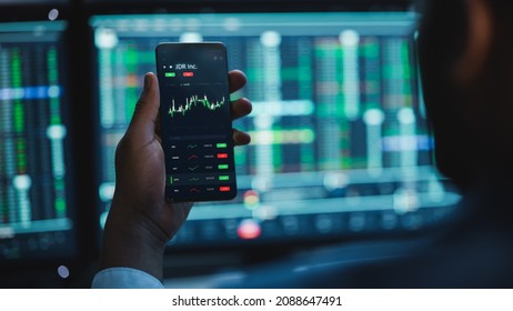 Financial Analyst Using Smartphone With Real-Time Stocks, Commodities And Market Charts. Working On Multi-Monitor Workstation With Stock Charts. Businessman Works In Investment Bank In The Evening.